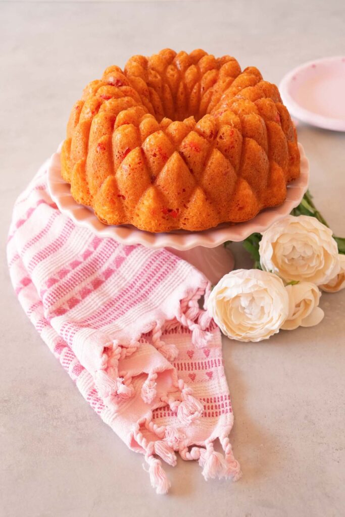 A strawberry bundt cake on a marble surface with a pink napkin and white peonies.