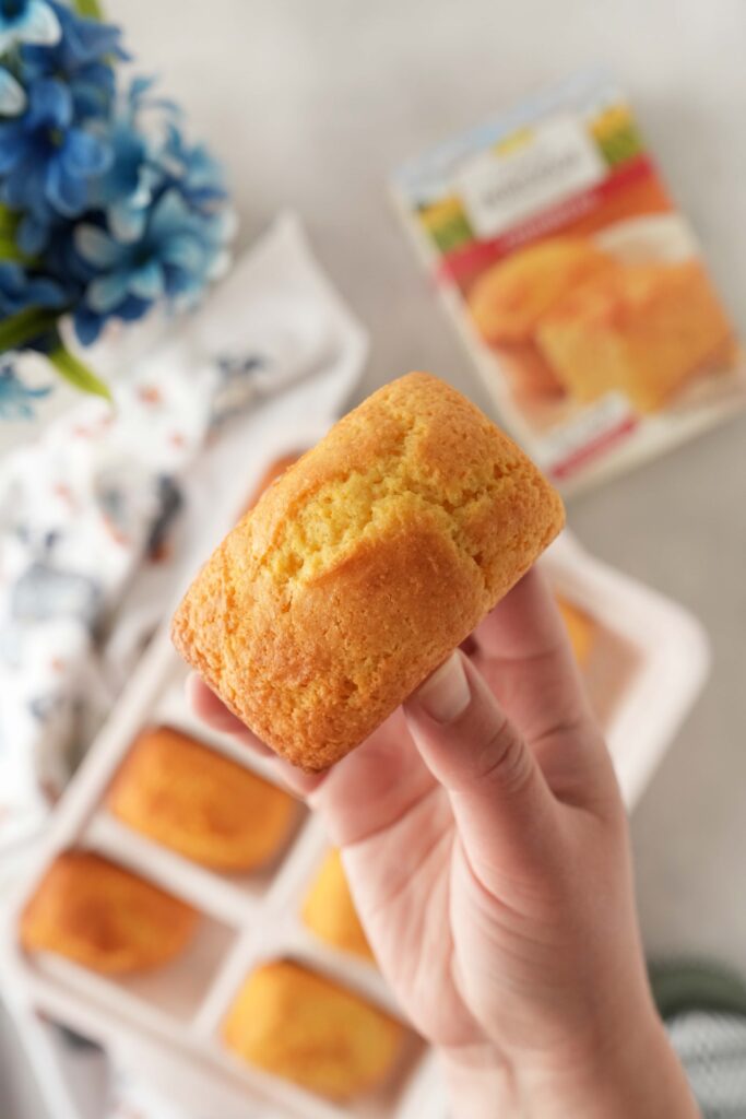 A hand holding up a mini cornbread above a blue floral arrangement and box of cornbread mix.