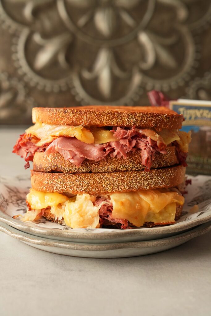 A double-stacked corned beef and cheese sandwich on a stone surface with a metal fleurs de les sign in the background.