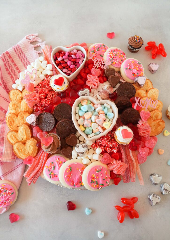 A valentine's day board with colorful pink, and red candies, baked goods, and conversation hearts.