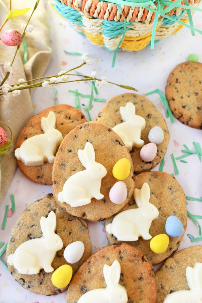 Several egg shaped chocolate chip cookies with a white chocolate bunny and pastel colored candy shelled chocolate eggs decorating the tops.