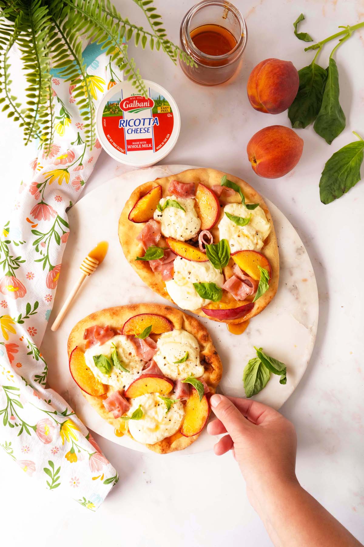 Woman's hand holding the edge of a naan flatbread baked and topped with ricotta cheese mounds, peach slices, and drizzled with hot honey. Setting is a white marble board on a granite countertop.