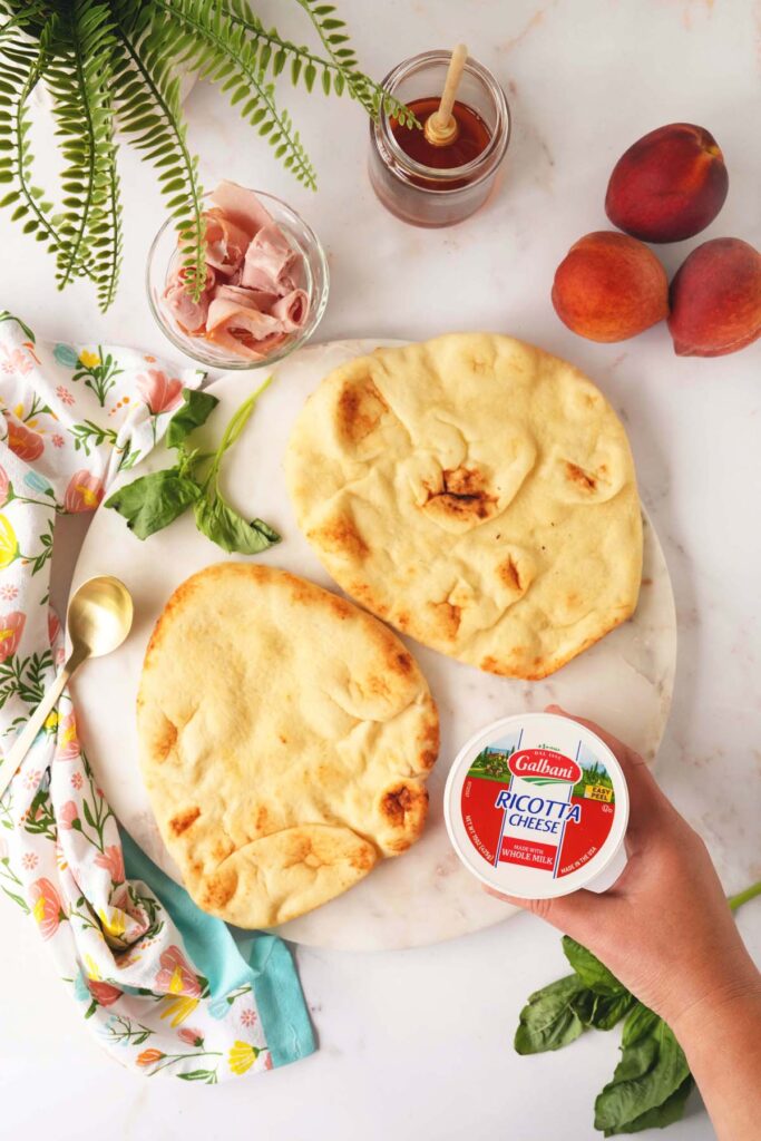 Woman's hand holding a container of Gilbani Ricotta Cheese. Two naan bread pieces, ham, honey, and peaches on a countertop.